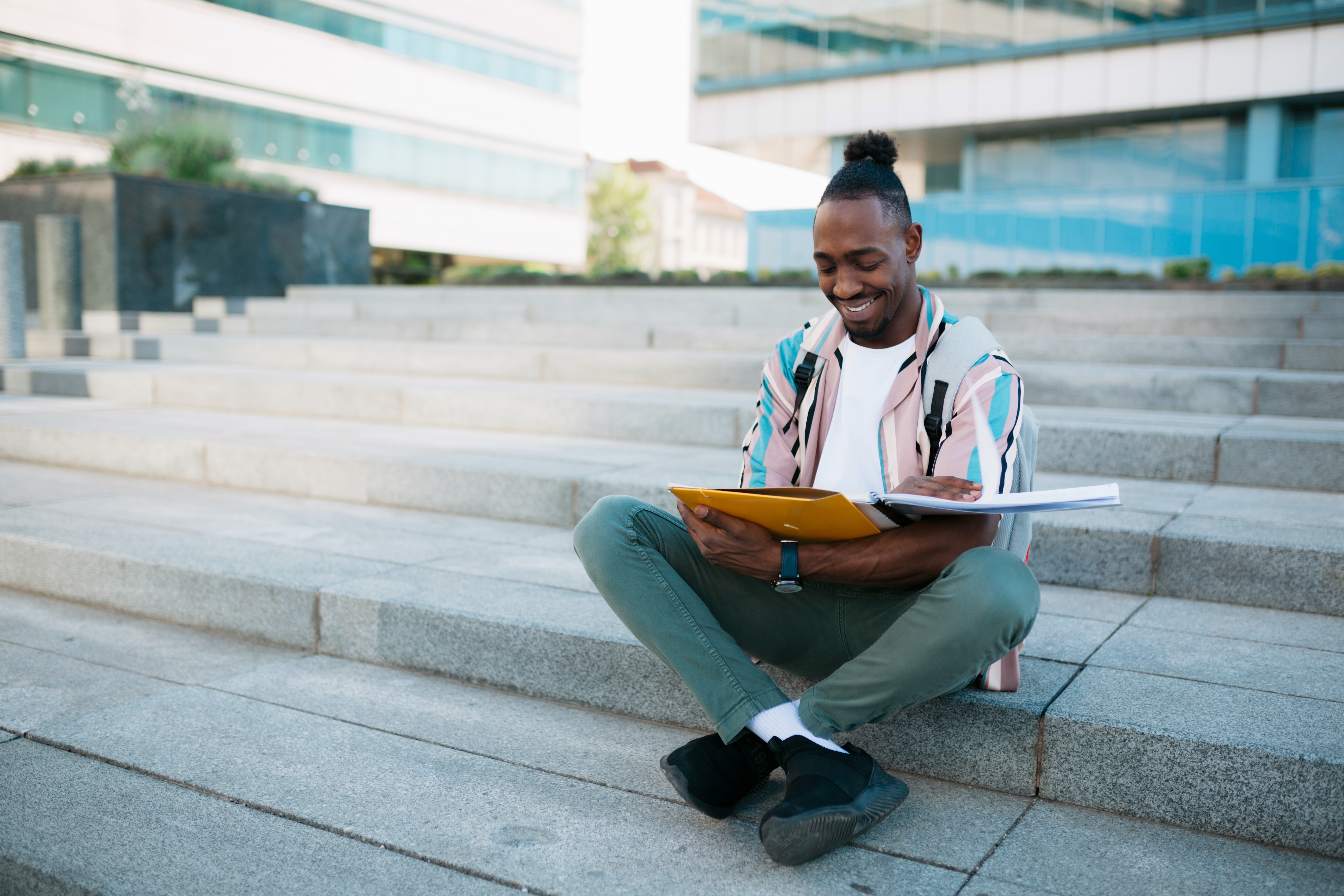 Person sitting on outdoor steps reading a book and holding a yellow folder, with a modern building in the background - Pearson TalentLens